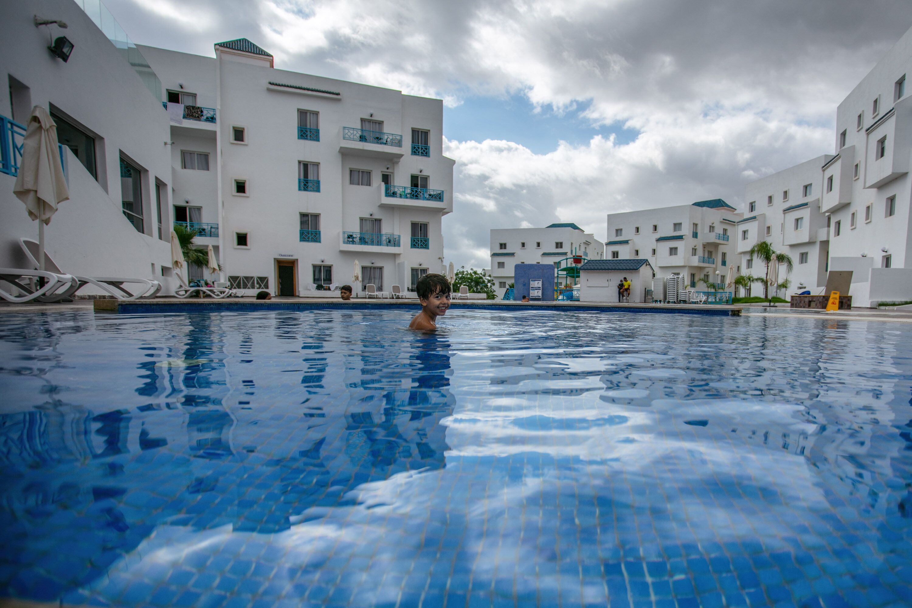 A swimming pool with clear blue water, surrounded by white buildings with balconies and blue railings.