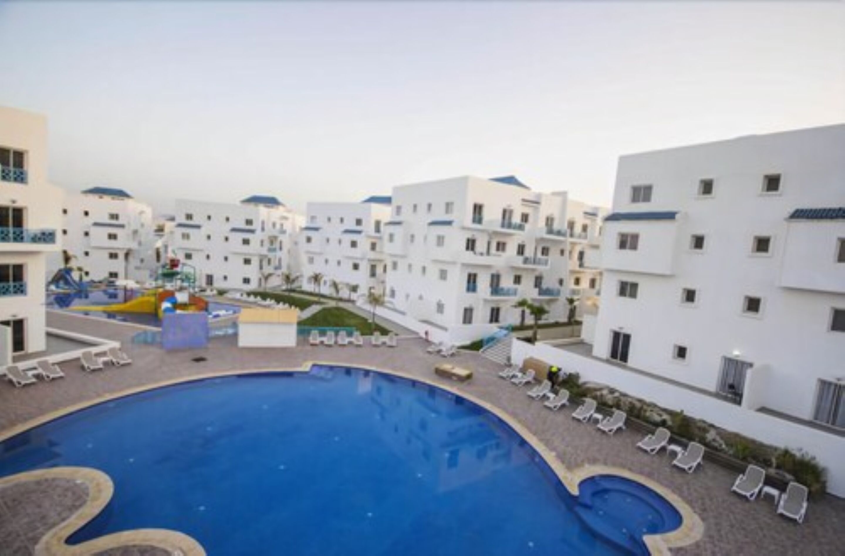 A resort with a large swimming pool, multiple white buildings, and sun loungers.