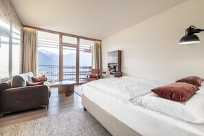 A modern hotel room with a large bed, a sofa, a small table, and a view of mountains through large windows. - NIDUM - Casual Luxury Hotel (Telfs)