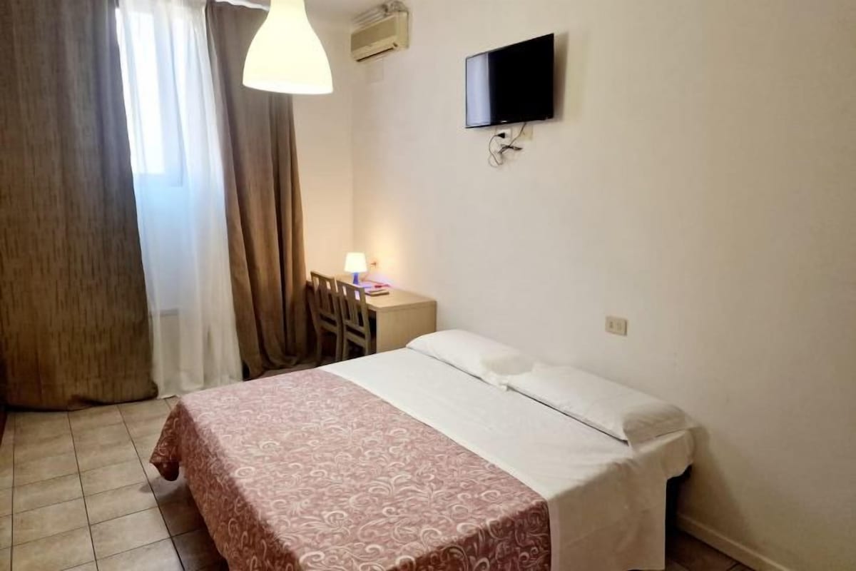 A hotel room with a bed, a desk, a chair, a TV mounted on the wall, and a window with curtains.