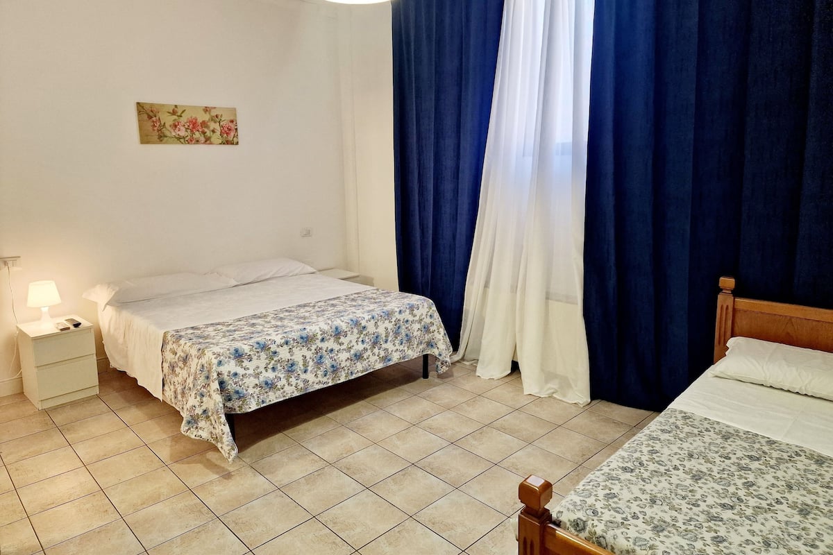 A hotel room with two beds, a nightstand with a lamp, a wall clock, and a window with blue curtains.