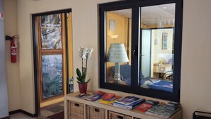 A hallway with a fire extinguisher, a window with a view of a building, a potted orchid, a lamp, and a table with books.