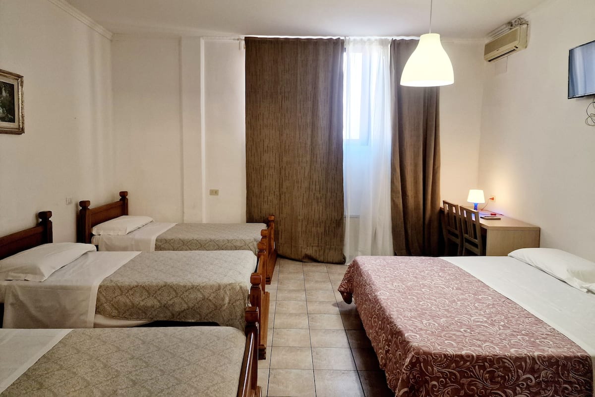 A hotel room with two single beds, a large bed, a desk with a lamp, and a TV on the wall.