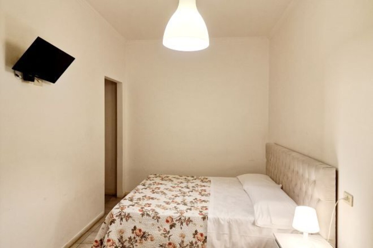 A small, neatly arranged bedroom with a bed, a bedside lamp, and a wall-mounted TV.
