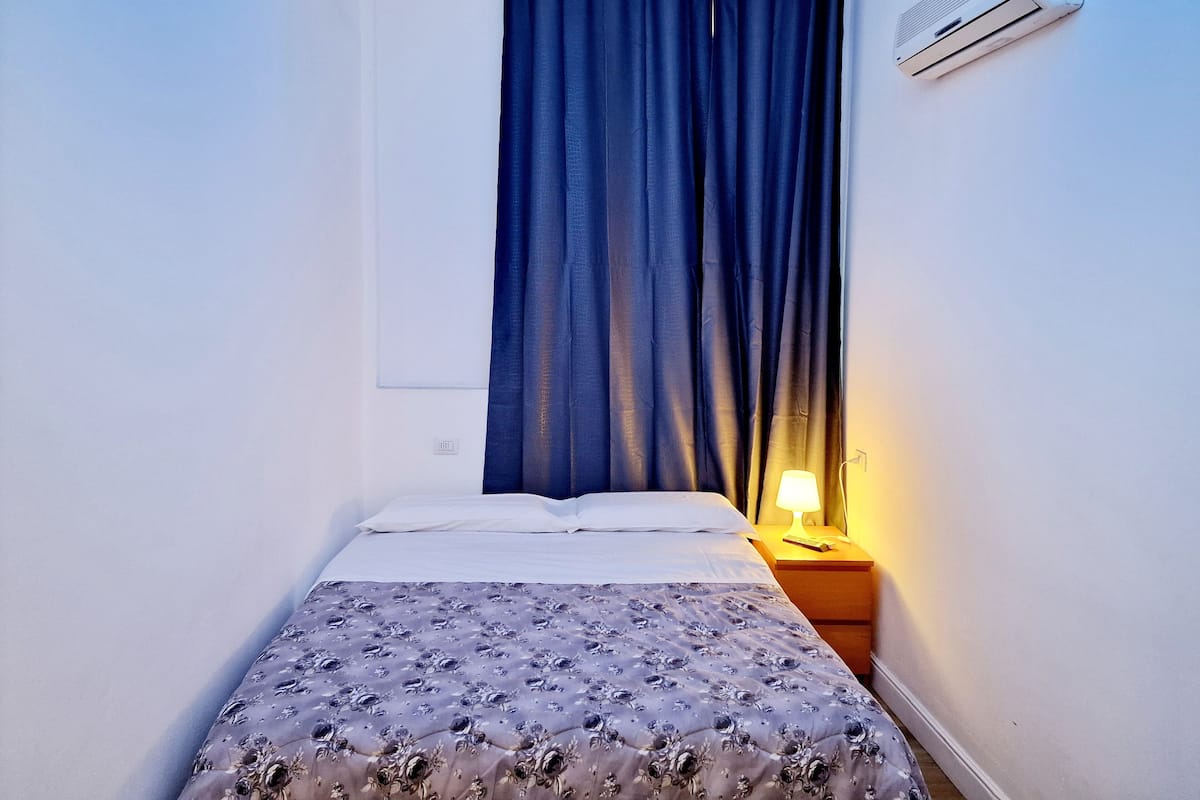 A small, single-bed room with a bed, a nightstand with a lamp, a window with curtains, and an air conditioning unit on the wall.