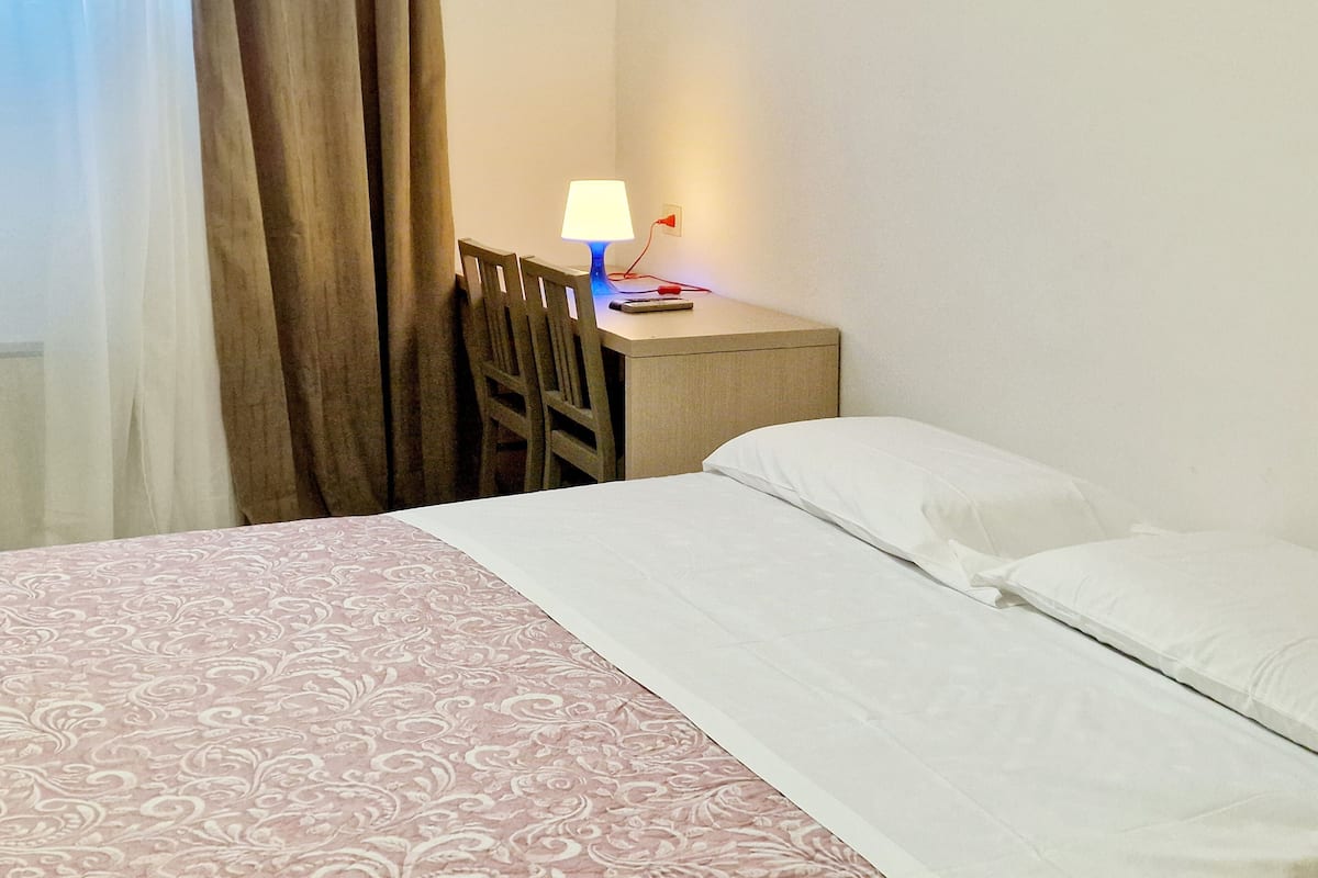 A hotel room with a bed, a desk with a lamp, a chair, and a window with curtains.