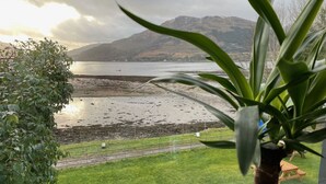 A view of a beach with mountains in the background, a grassy area in the foreground, and a potted plant in the foreground. - The Shore House (Cairndow)
