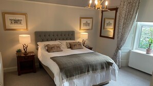A neatly arranged bedroom with a tufted headboard, a large bed with multiple pillows, bedside lamps, framed pictures on the walls, and a window with a view of greenery. - The Shore House (Cairndow)