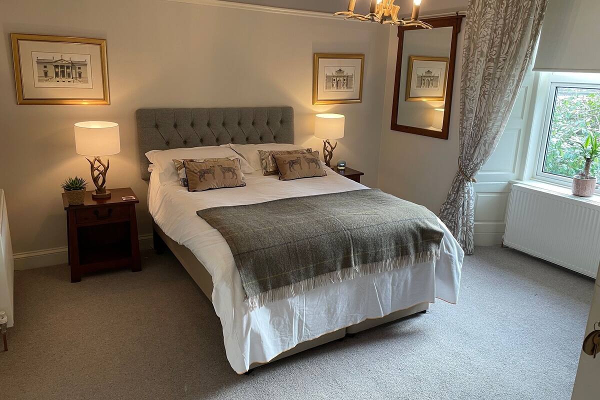 A neatly arranged bedroom with a tufted headboard, a large bed with multiple pillows, bedside lamps, framed pictures on the walls, and a window with a view of greenery.