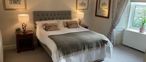 Een netjes ingerichte slaapkamer met een gestoffeerd hoofdbord, een groot bed met veel kussens, nachtlampjes, ingelijste foto’s aan de muur en een raam met uitzicht op groen.