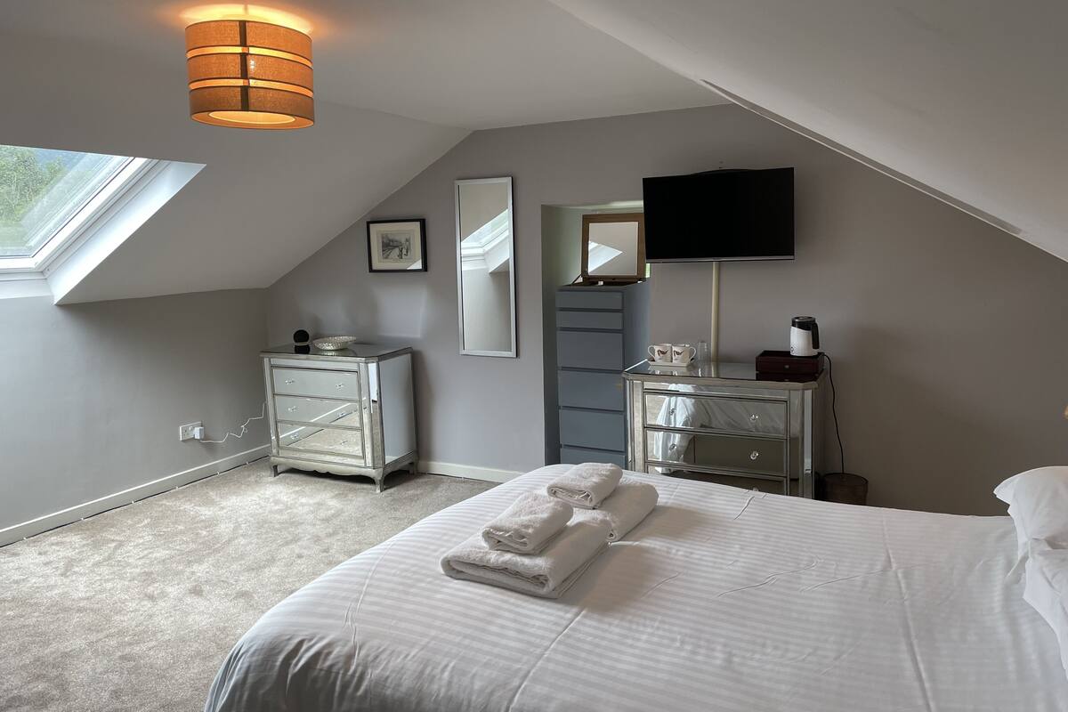 A bedroom with a sloped ceiling, a bed with white linens, a dresser, a vanity with a mirror, and a flat-screen TV mounted on the wall.