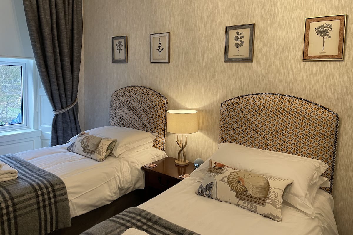 A hotel room with two beds, a nightstand with a lamp, and three framed botanical prints on the wall.