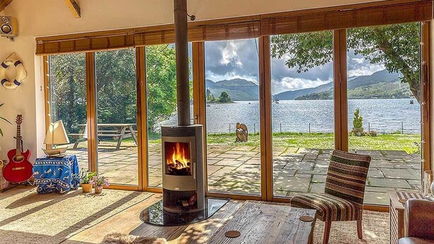A cozy room with a wood-burning stove, large windows, a guitar, and a view of a lake and mountains. - The Shore House (Cairndow)