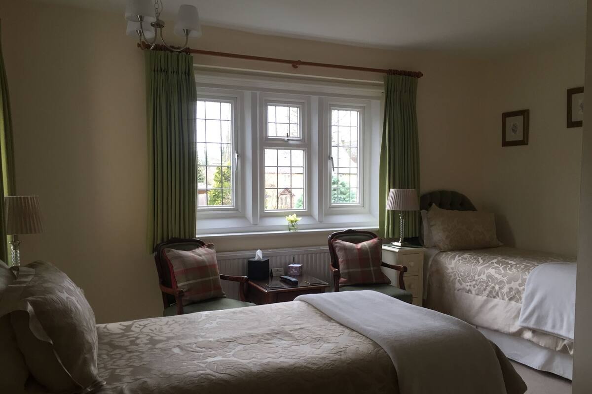 A hotel room with two beds, a window with curtains, a bedside table with a lamp, and a chair with a pillow.