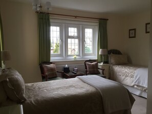 A hotel room with two beds, a window with curtains, a bedside table with a lamp, and a chair with a pillow. - Coombe House (Cheltenham)