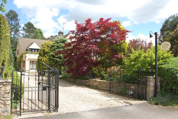 A gated entrance to a property with a stone wall and a metal gate, surrounded by lush greenery and trees with red leaves. - Coombe House (Cheltenham)