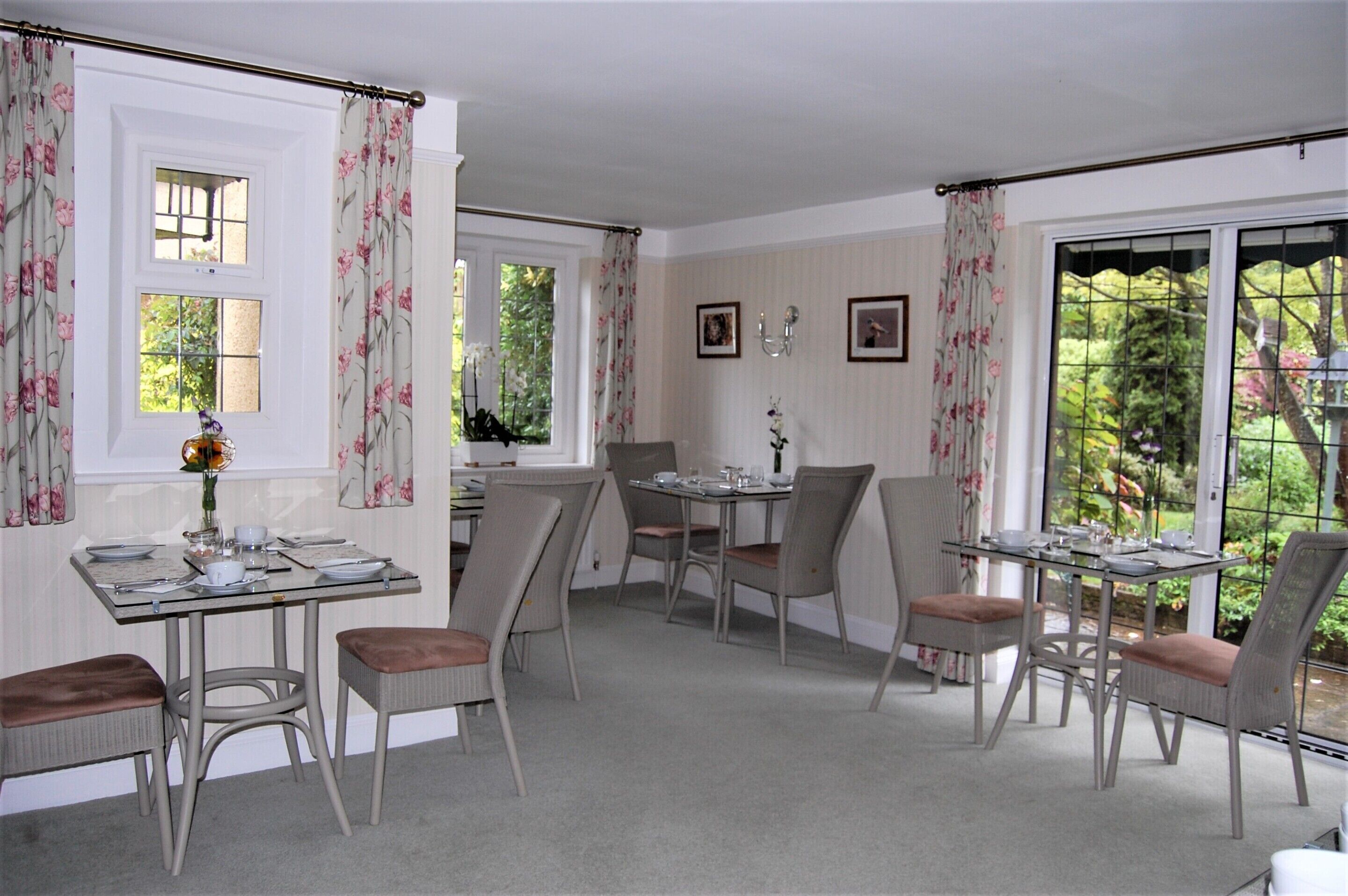 A dining area with two tables, each set for four. The room has large windows with curtains, a view of greenery outside, and a wall-mounted light fixture.