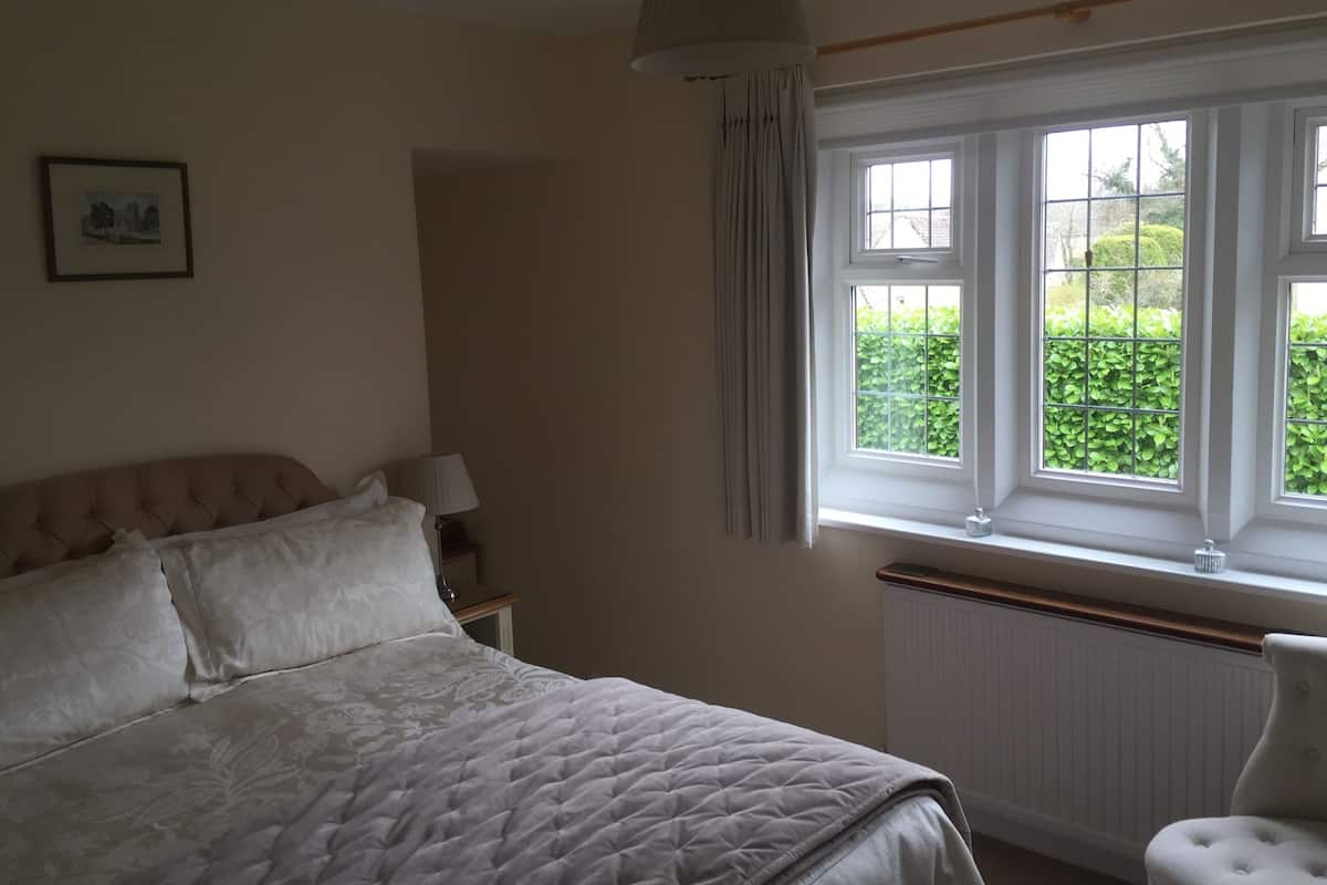 A bedroom with a bed, a nightstand, a lamp, a window with a view of greenery, and a radiator.