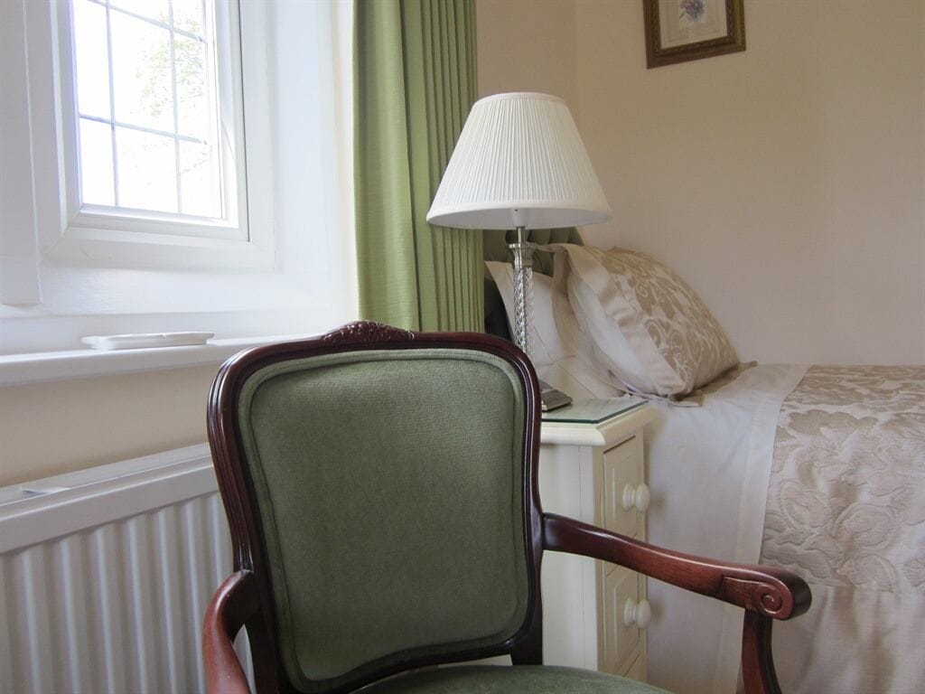A room with a wooden chair, a bedside table with a lamp, a bed with a patterned bedspread, and a window with curtains.