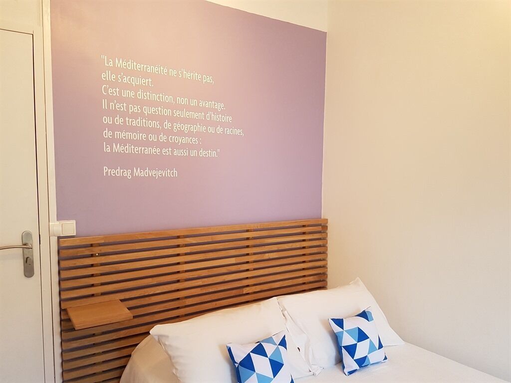 A bedroom with a wooden headboard, a bed with white bedding and blue patterned pillows, and a wall with a quote in French.