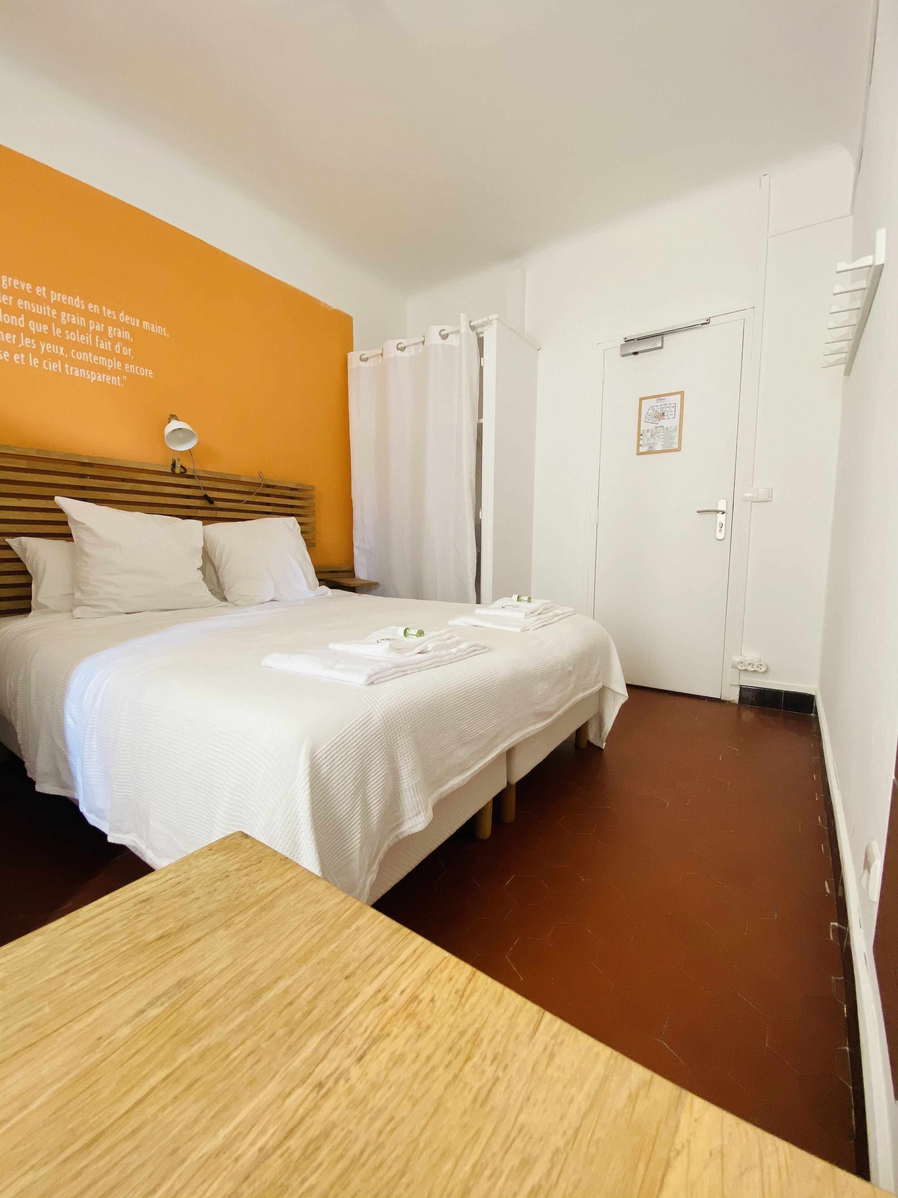 Une chambre d’hôtel avec un lit, une table en bois, un mur blanc orné d’une citation, une porte blanche et un plancher marron.