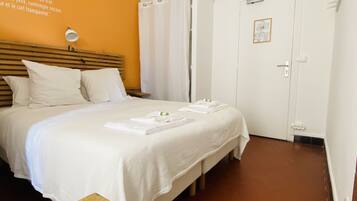 A hotel room with a bed, a wooden table, a white wall with a quote, a white door, and a brown floor.