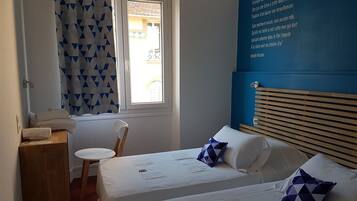 Un dormitorio con dos camas, cabeceros de madera, una ventana con cortina geométrica y una pared con una cita.