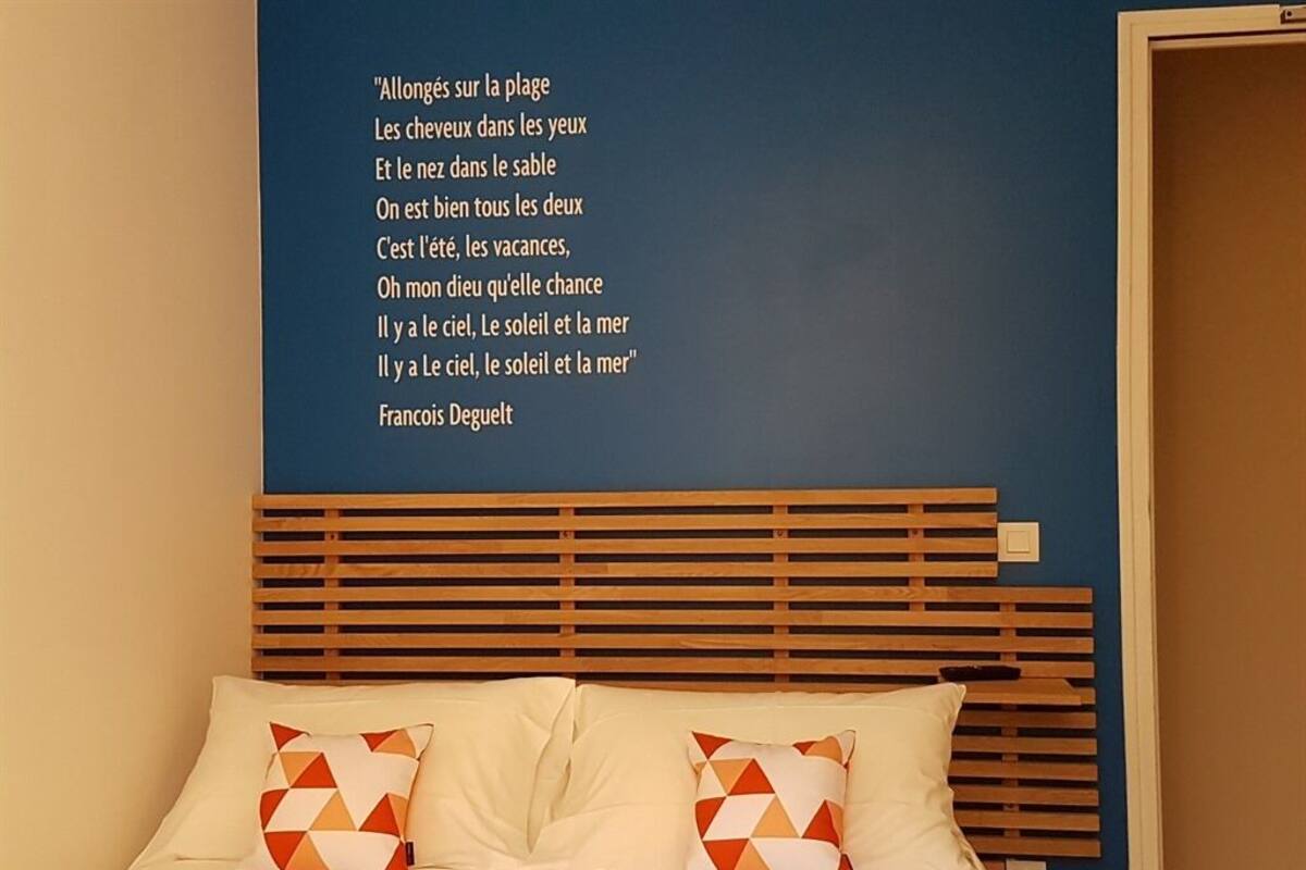 Un dormitorio con cama, almohadas, cabecera de madera y una pared con un poema francés.