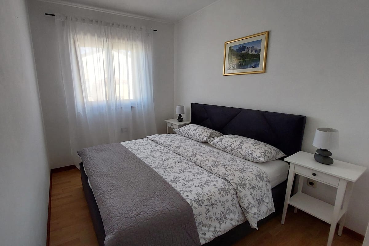 A bedroom with a bed, a nightstand, a lamp, a window with curtains, and a framed picture on the wall.
