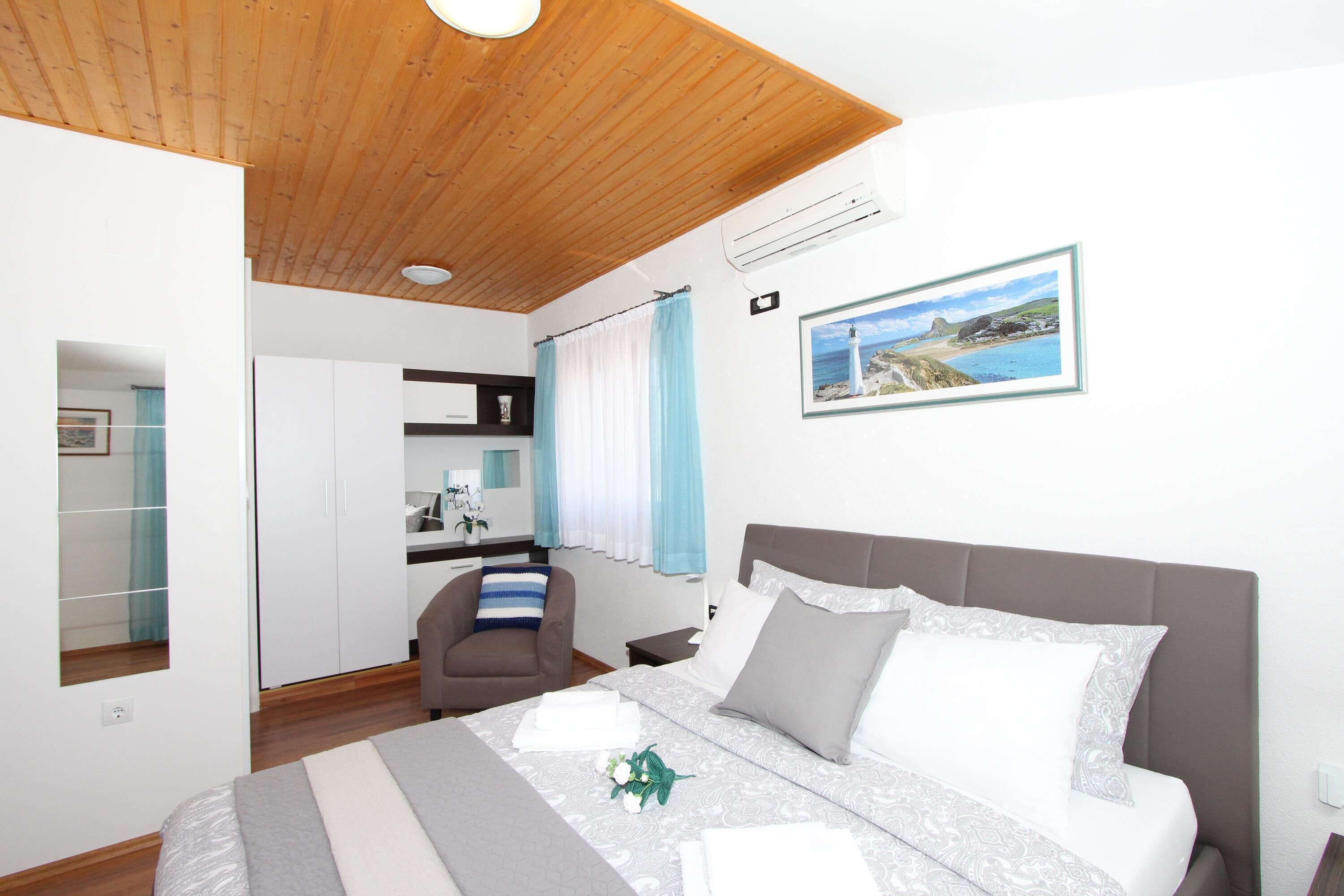 A bedroom with a wooden ceiling, a bed with grey and white bedding, a chair, a nightstand, and a picture of a coastal scene.
