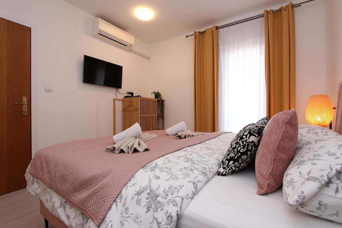 A hotel room with a bed, a TV mounted on the wall, an air conditioning unit, and a window with curtains.