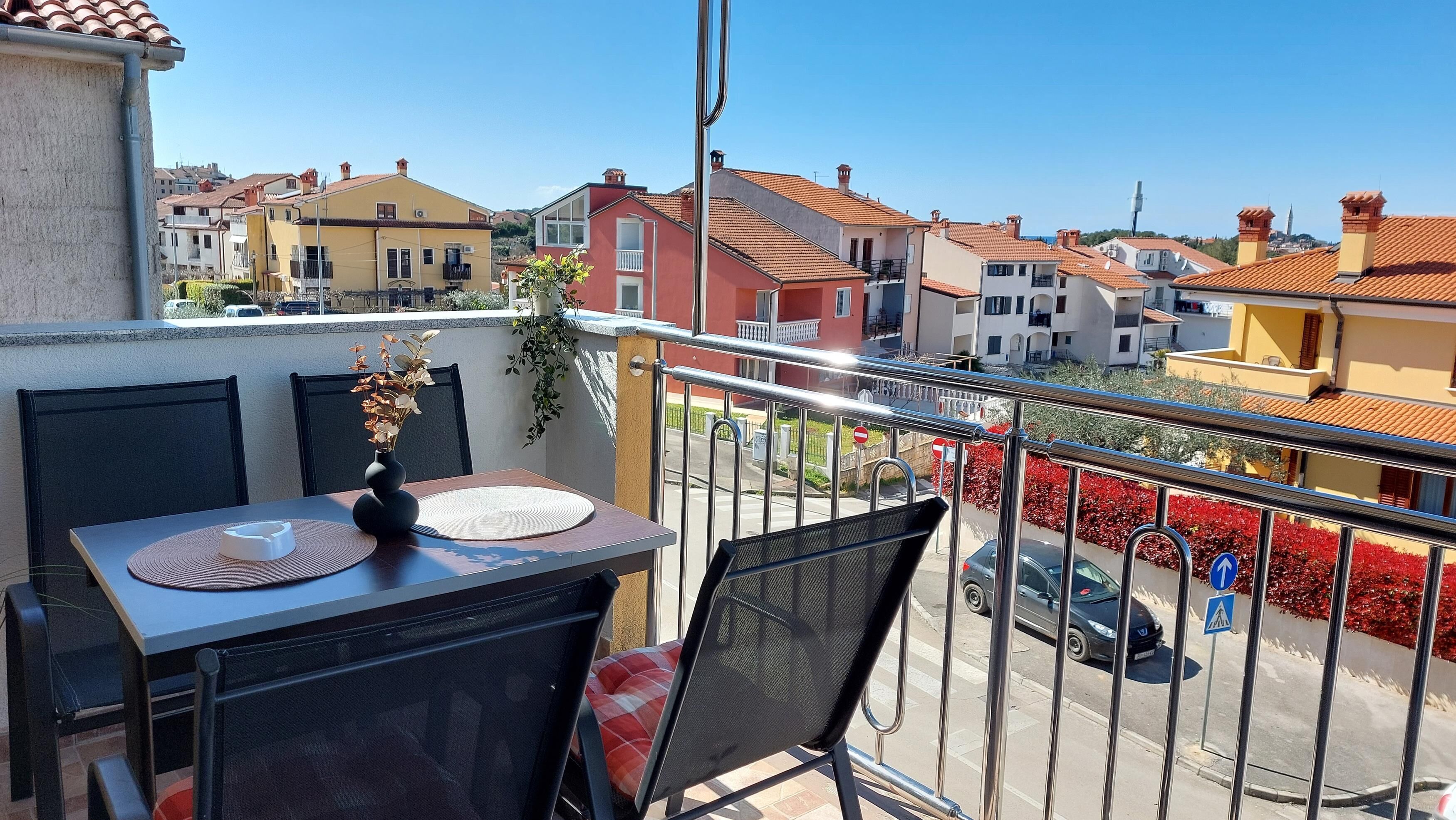 A balcony with a table and chairs, overlooking a residential area with colorful houses and a clear sky.