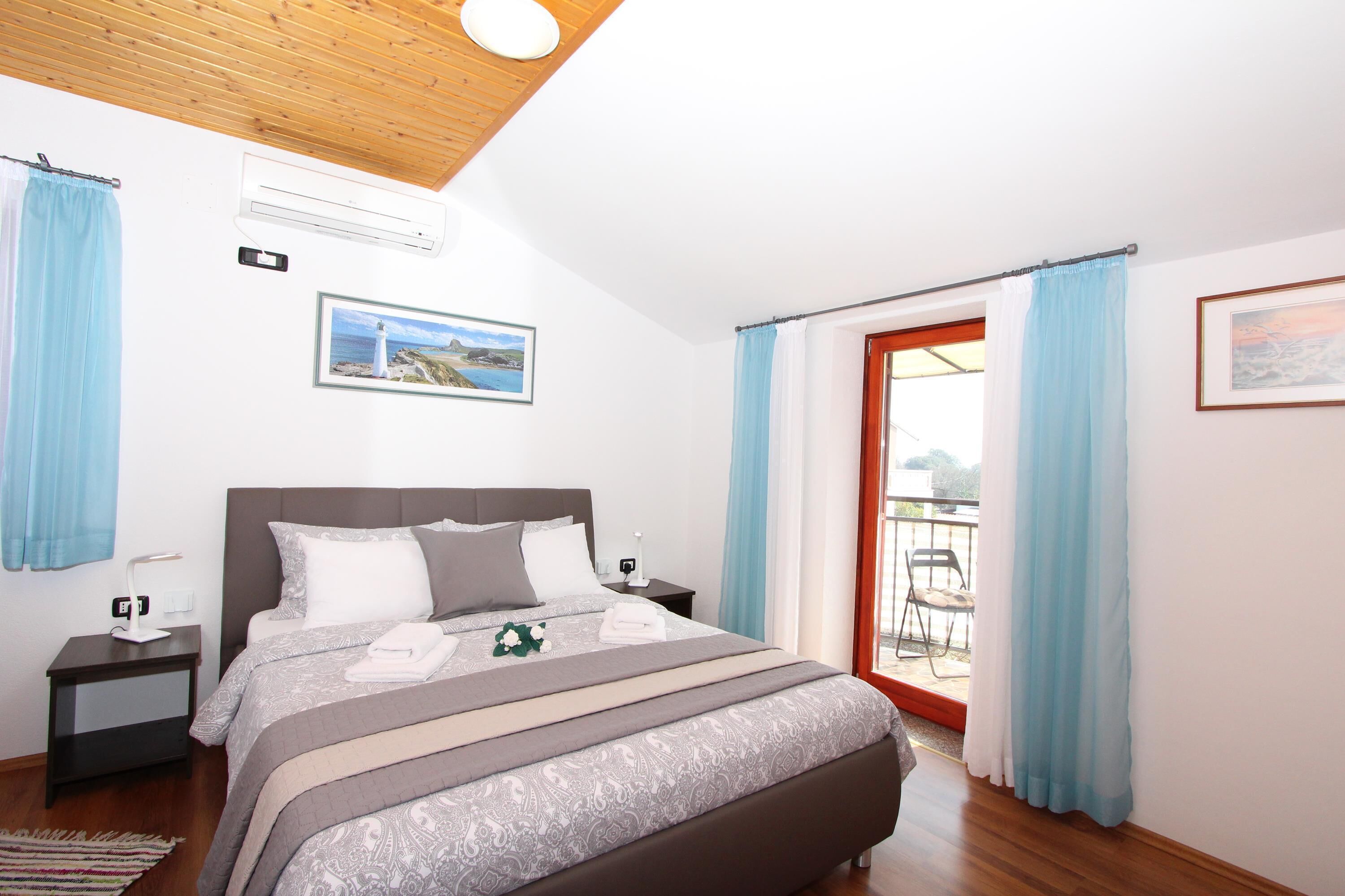 A bedroom with a bed, nightstands, an air conditioner, a picture on the wall, and a view through a door.
