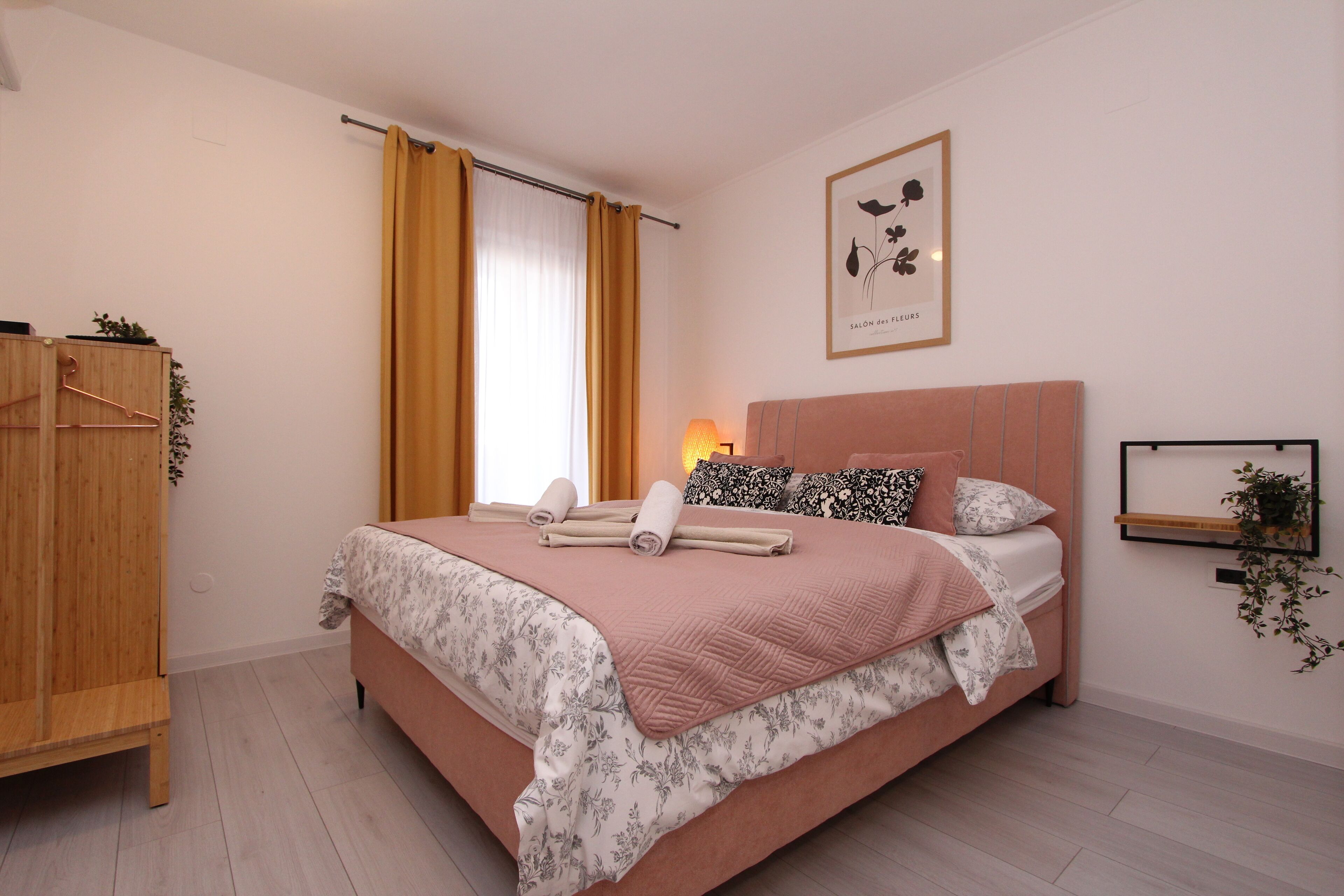 A neatly made bed with a pink headboard, a wooden wardrobe, and a framed picture on the wall.