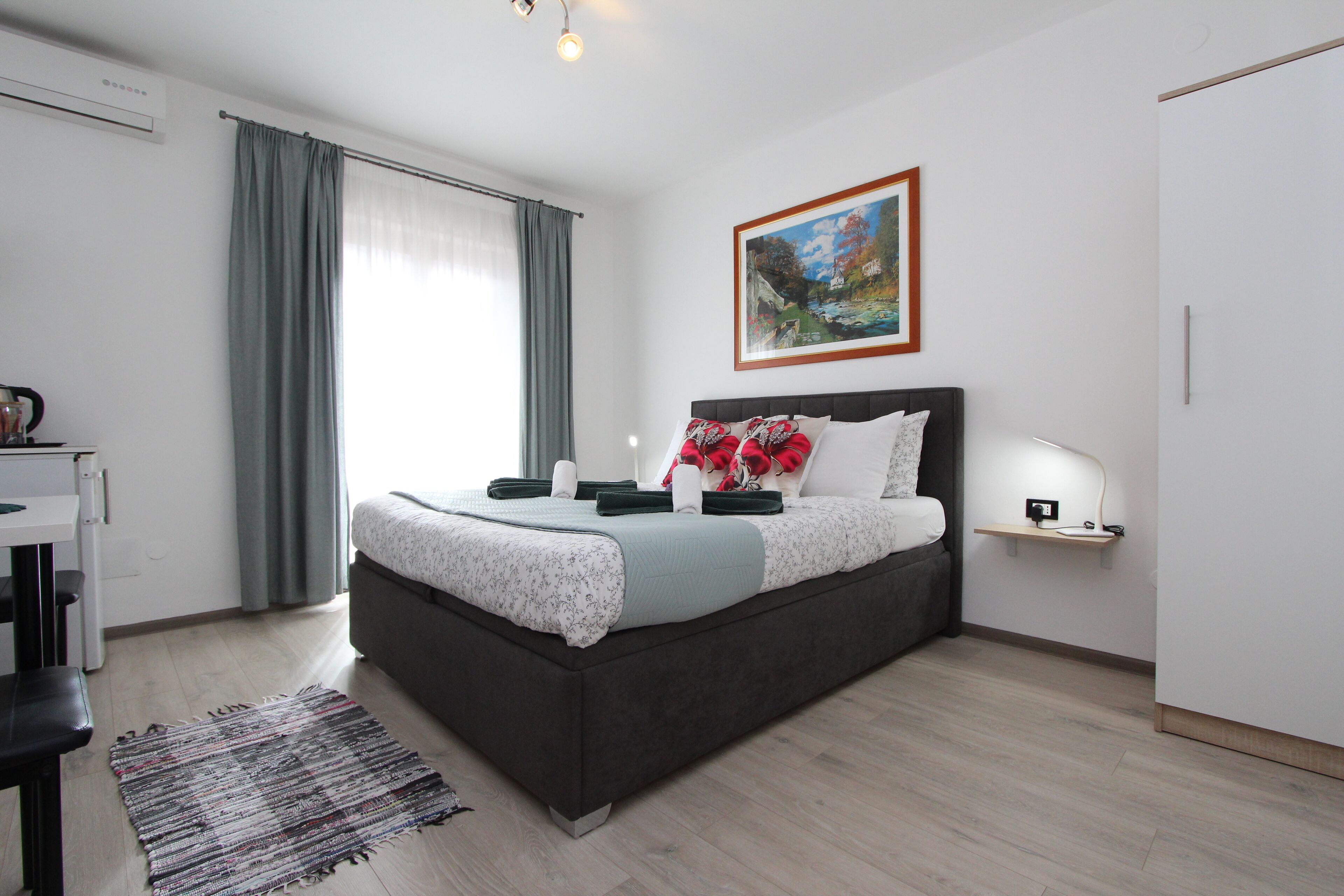 A modern bedroom with a dark headboard bed, a bedside table with a lamp, a painting on the wall, and a large window with curtains.