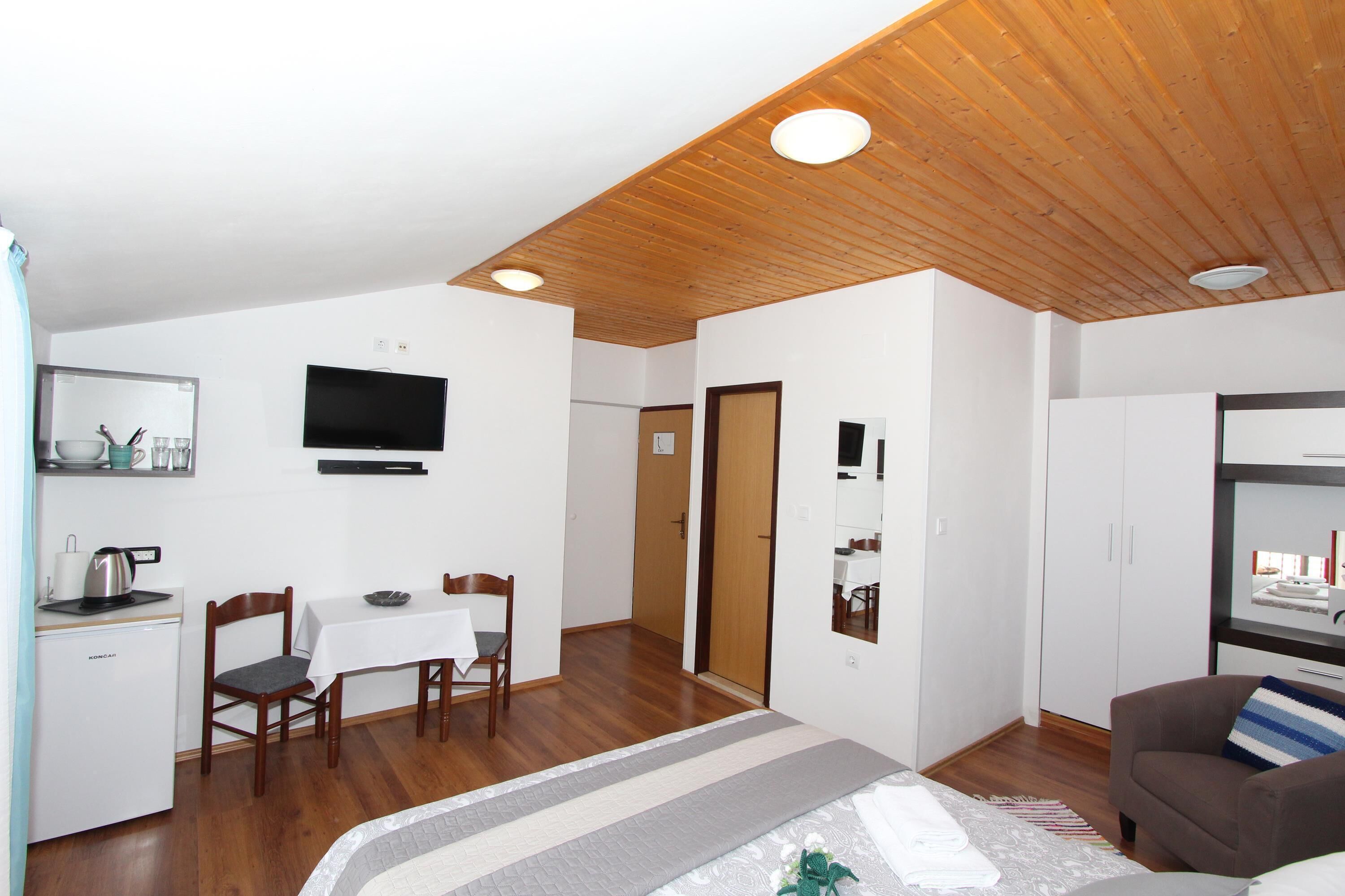 A compact room with a bed, a dining table with chairs, a kitchenette, a TV mounted on the wall, and a wooden ceiling.