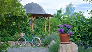 A gazebo surrounded by lush greenery, a bicycle, and a potted plant with purple flowers. - Babiččina Zahrada Penzion & Restaurace (Pruhonice)