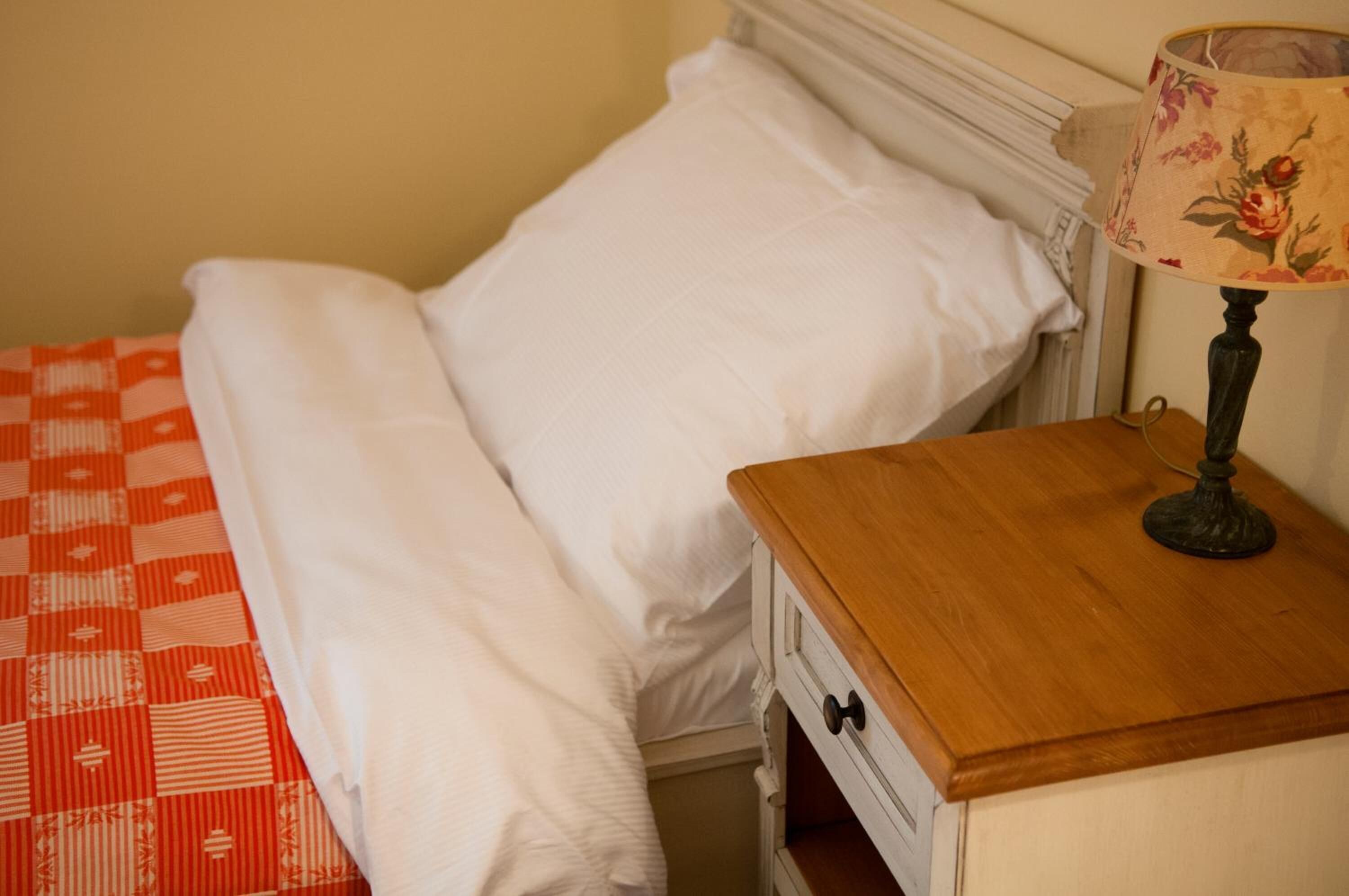 A neatly made bed with white linens, a wooden bedside table with a lamp, and a patterned red and white blanket.