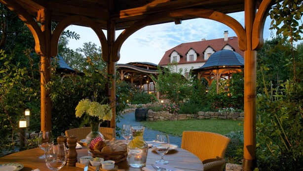 A wooden gazebo with a table set for dining, surrounded by lush greenery and a stone wall. - Babiččina Zahrada Penzion & Restaurace (Pruhonice)