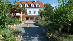 A white building with a red roof, surrounded by a garden with a gravel path leading to the entrance. - Babiččina Zahrada Penzion & Restaurace (Pruhonice)