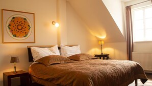A bedroom with a large bed, a nightstand with a lamp, a wall-mounted light, and a window with curtains. - Babiččina Zahrada Penzion & Restaurace (Pruhonice)