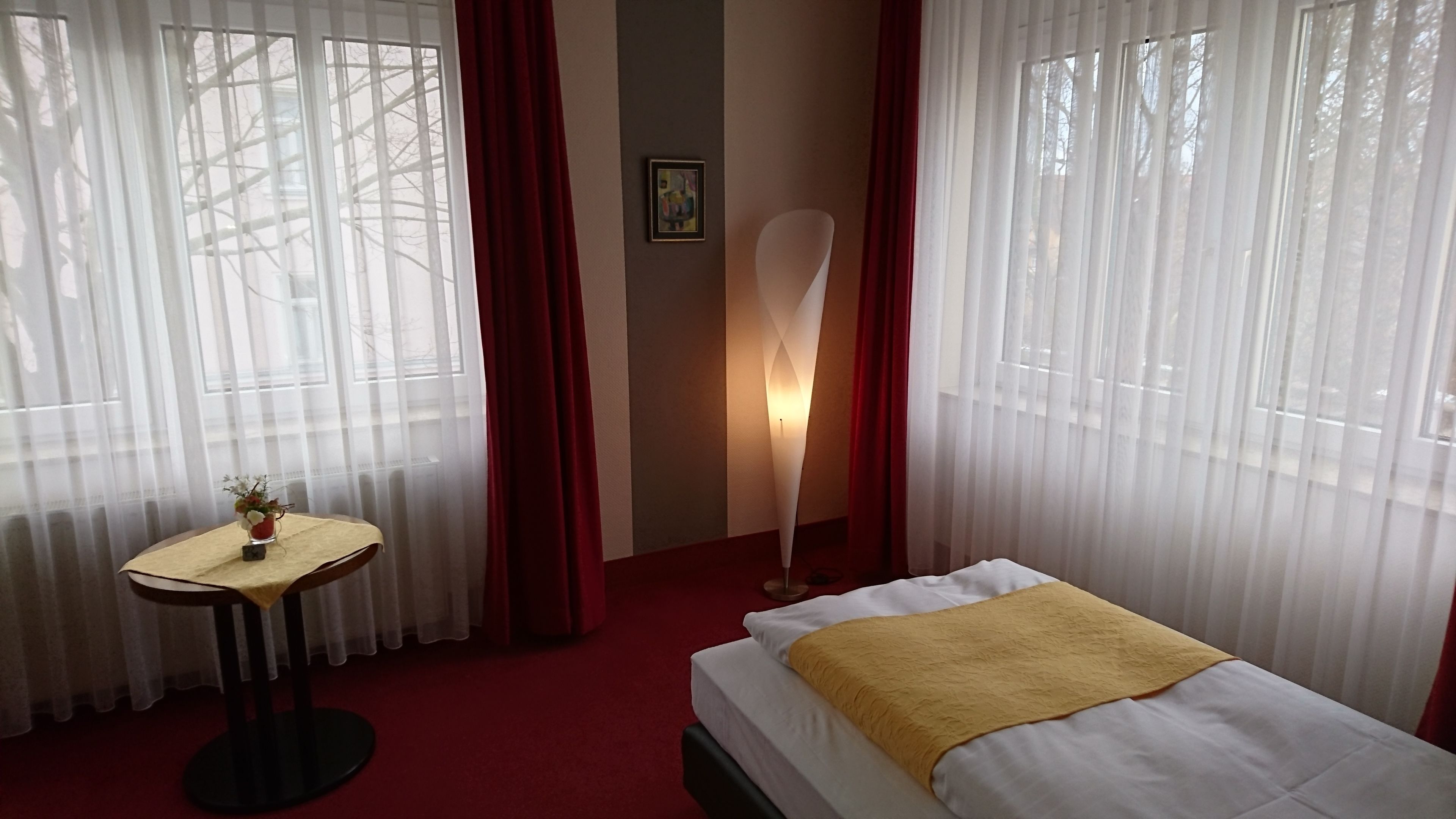 A hotel room with a bed, a small table with a vase, a standing lamp, and sheer curtains.