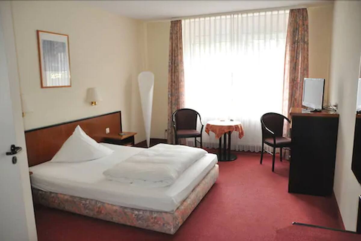A hotel room with a bed, a desk with a computer, two chairs, a small table, and a window with curtains.