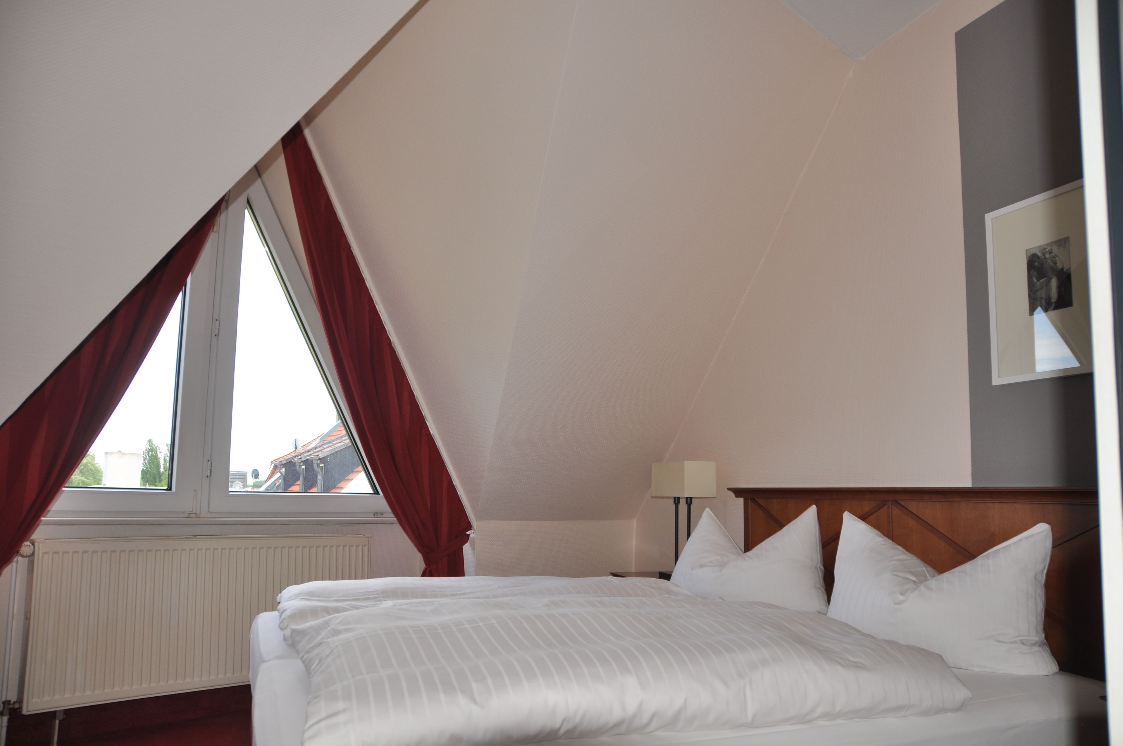 A bedroom with a sloped ceiling, a bed with white linens, a nightstand with a lamp, and a window with red curtains.