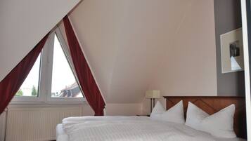 A bedroom with a sloped ceiling, a bed with white linens, a nightstand with a lamp, and a window with red curtains.