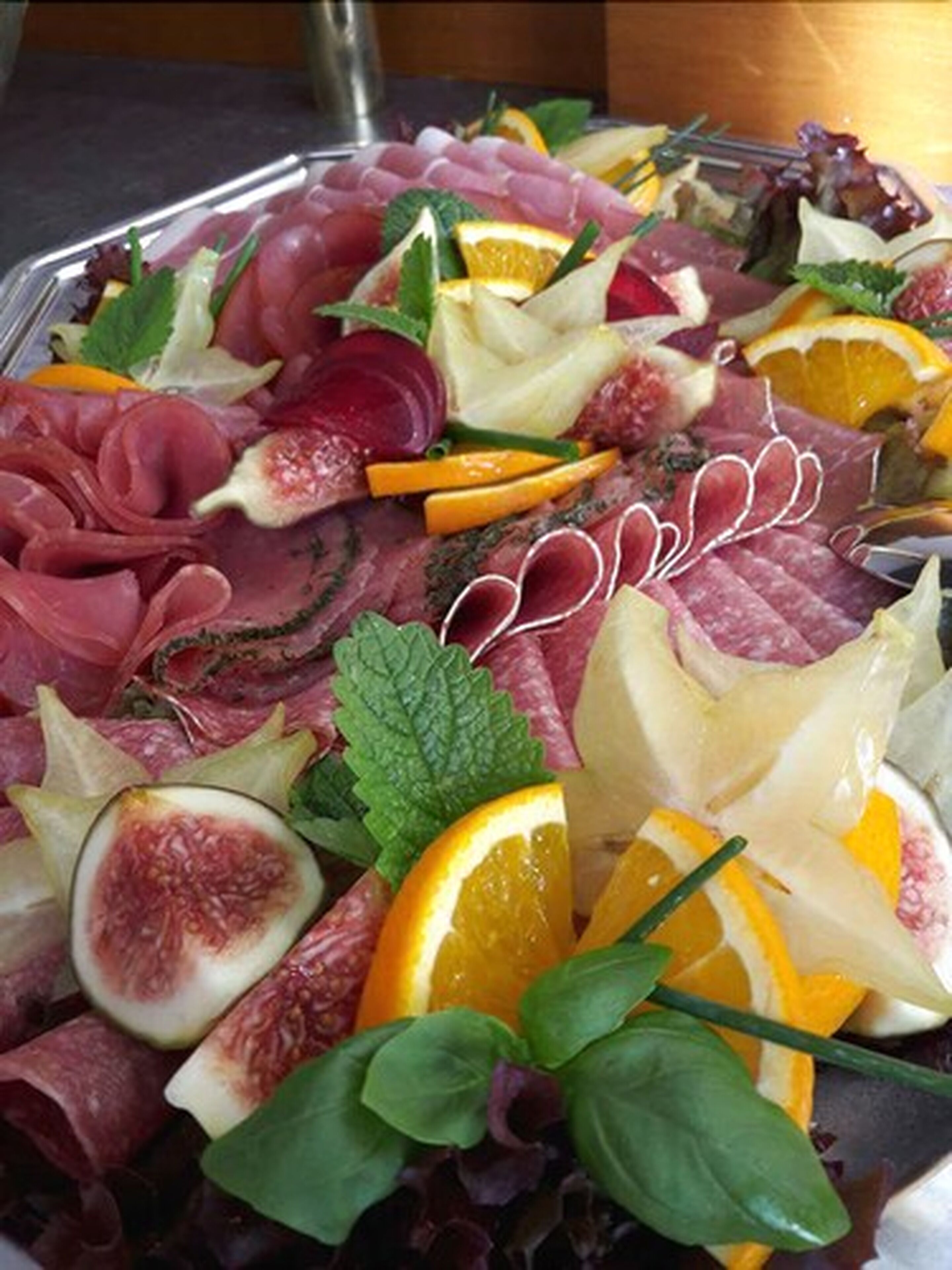 A platter of assorted meats and fruits, including prosciutto, figs, and oranges, garnished with herbs.