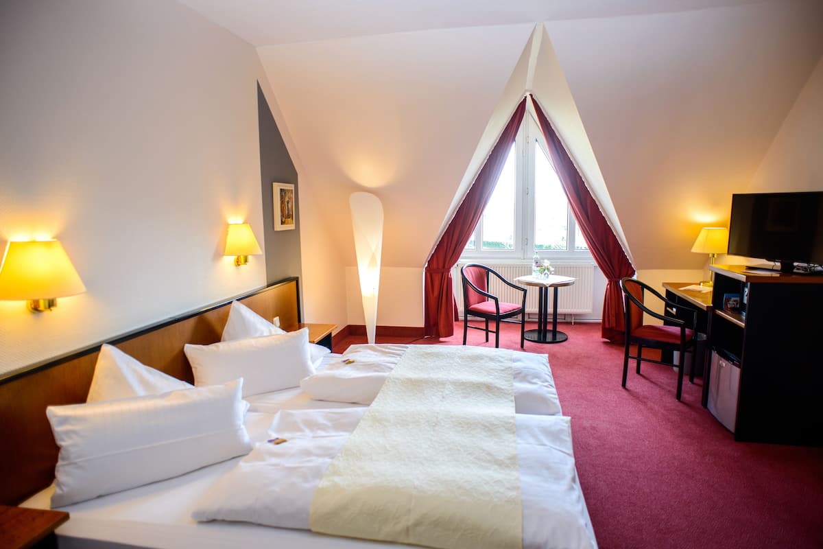A hotel room with a large bed, two bedside tables, a desk with a chair, a TV, and a window with curtains.
