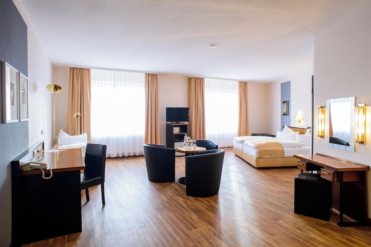 A modern hotel room with a wooden floor, a bed, a desk, a chair, a TV, and a large window with curtains.