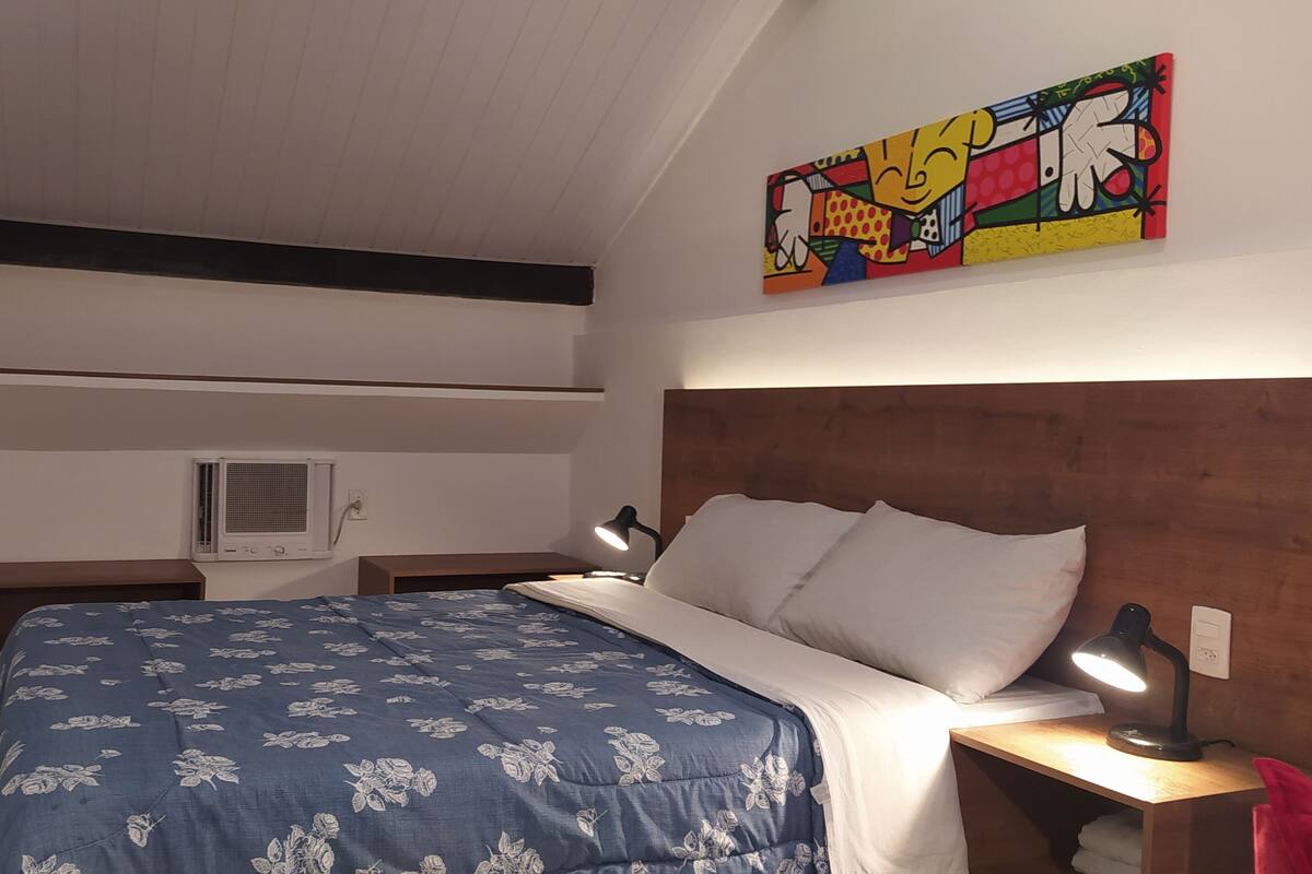 A bedroom with a bed, bedside lamps, a wall-mounted air conditioner, and a colorful artwork above the bed.
