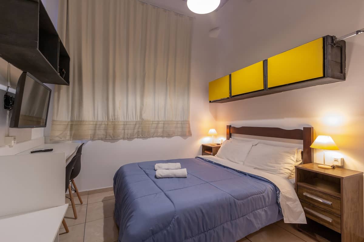 A hotel room with a bed, bedside tables, a desk, a chair, a TV, and a window with curtains.